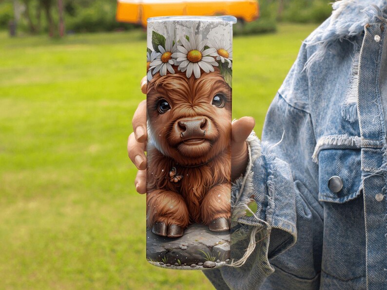 May include: A stainless steel tumbler featuring a detailed illustration of a Highland calf with a daisy flower crown. The calf is brown with large eyes and a small butterfly. The tumbler is held against a green background.