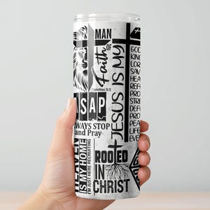 May include: A white tumbler with black text and graphics. The design includes the words "Faith," "Jesus is My," and "Rooted in Christ," along with a lion illustration and a cross. The tumbler is held by a hand.