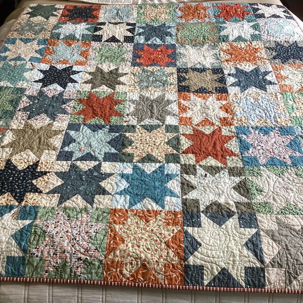 May include: A patchwork quilt featuring a repeating star pattern in various colors including rust orange, navy blue, sage green, and cream. The fabric squares are stitched together with intricate quilting lines throughout the entire surface.