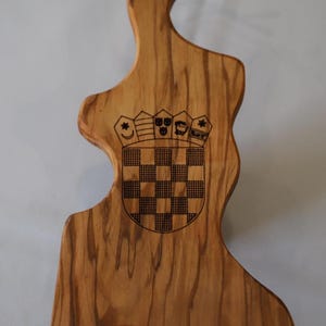 Puede incluir: Tabla de cortar de madera con una forma única y un emblema decorativo. La tabla es de madera marrón claro y presenta un diseño de escudo de armas. Se adjunta un lazo de cuerda natural para colgar. La tabla mide aproximadamente 30 cm de largo.