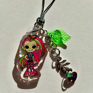 Peut inclure: Un charm en acrylique transparent représentant un personnage de dessin animé aux cheveux roses et verts, vêtu d'une tenue noire et de bottes roses. Le charm est relié à une chaîne argentée avec une feuille verte et un charm en forme de cœur aux facettes irisées.