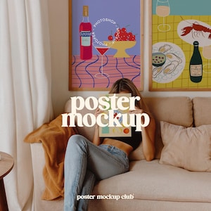 May include: A woman sitting on a sofa holding a poster mockup with the text "poster mockup" in front of her face. The poster is a colourful illustration of a glass of wine, cherries, and a bottle of Campari. The sofa is beige with a brown throw blanket and a beige cushion.