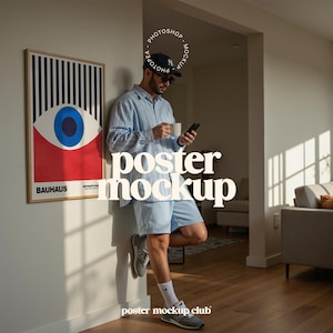 May include: A framed Bauhaus art print with a striking eye design in red, blue, and white hangs on a wall. A person in a blue shirt and shorts holds a phone and a mug. The words "poster mockup" are displayed. The image is set in a domestic interior.
