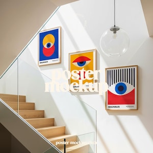 Może przedstawiać: Trzy oprawione plakaty z grafiką Bauhaus z geometrycznymi wzorami oczu w kolorach czerwonym, niebieskim, żółtym i czarnym wiszą na białej ścianie. Plakaty są w drewnianych ramach. Widoczne są również szklane schody i przezroczysta oprawa oświetleniowa.