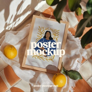 Puede incluir: Un marco de madera con una maqueta de póster en su interior. El póster presenta a una mujer con cabello castaño y una camisa azul de pie frente a un diseño floral amarillo y naranja. El texto "poster mockup" está escrito en letras blancas en el póster. El marco está sentado sobre un paño blanco con un limón a la izquierda del marco y otro limón a la derecha del marco.