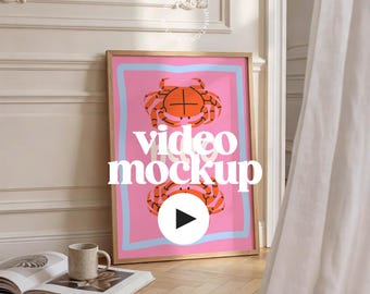 May include: A framed poster featuring a pink background with two orange crab illustrations. The poster has a light blue border and the words "video mockup" in white. A coffee cup and an open book are on the floor.