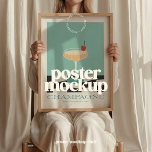 May include: A poster mockup with a wooden frame, featuring a glass of champagne with a cherry on top. The poster has the text "poster mockup" and "CHAMPAGNE" in a retro style font.