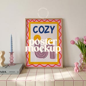 Könnte beinhalten: Ein rechteckiges gerahmtes Poster mit dem Wort COZY in blauen Buchstaben über einer Grafik von zwei Kerzen mit gelben Flammen und rosa gestreiften Körpern. Das Poster hat einen gelben Innenrand und einen rosa gewellten Außenrand auf weißem Grund.