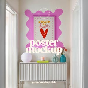 May include: A white cabinet with a fluted front, a white lamp, a candle, a book, a blue vase with flowers, and a framed poster with the text "you're in my heart" on a white wall with a pink splatter paint design.