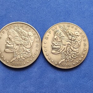 Puede incluir: Dos monedas de plata con un diseño de calavera, con las inscripciones "E PLURIBUS UNUM" y "LIBERTY". Una moneda está fechada en 1878, la otra en 1879. Las monedas están sobre una superficie azul.