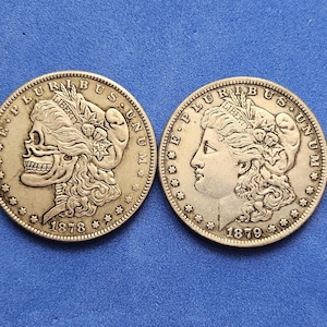Puede incluir: Dos monedas de plata Morgan. Una moneda presenta un diseño de calavera con la fecha de 1878, y la otra tiene el perfil de una mujer con la fecha de 1879. Ambas monedas tienen las palabras "E PLURIBUS UNUM" y "LIBERTY".