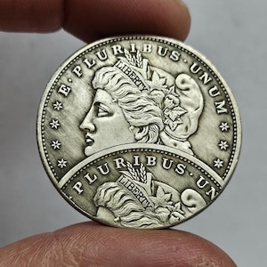 Puede incluir: Una moneda de plata con el perfil de una mujer, rodeada por las inscripciones "E PLURIBUS UNUM" y "LIBERTY". La moneda tiene un diseño detallado con estrellas y elementos florales.