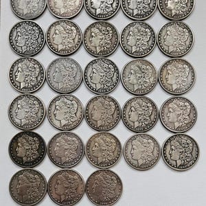 Puede incluir: Una colección de 30 monedas de plata de un dólar Morgan, cada una con una fecha diferente que va desde 1883 hasta 1903. Las monedas están dispuestas en filas, con las fechas visibles en cada moneda.