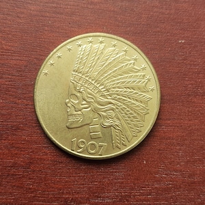 Puede incluir: Una moneda dorada con un diseño de tocado nativo americano con una cara de calavera. La palabra "LIBERTY" está inscrita, junto con el año "1907". La moneda tiene un diseño detallado en relieve y una forma circular.