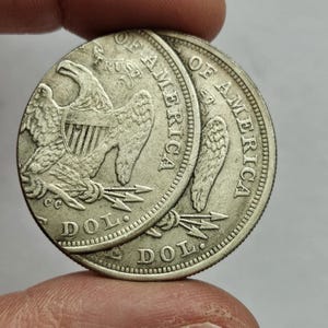Puede incluir: Dos monedas de plata, cada una con un águila con las alas extendidas, con las palabras "OF AMERICA" y "DOL." visibles. Las monedas se sostienen entre los dedos, mostrando su diseño detallado y su apariencia envejecida. Las monedas tienen un aspecto desgastado y antiguo.