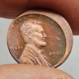 Puede incluir: Una moneda de centavo Lincoln de 1955 de color cobre. El anverso presenta un perfil de Abraham Lincoln, la palabra "LIBERTY" y la fecha "1955". La moneda también tiene la inscripción "IN GOD WE TRUST".