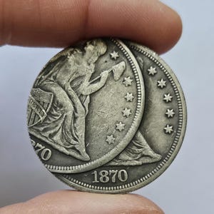 Könnte beinhalten: Eine Silbermünze von 1870 mit einer sitzenden Figur, die einen Stab und einen Schild hält, umgeben von Sternen. Die Münze ist teilweise verdeckt, wobei das Datum und das Wort "LIBERTY" sichtbar sind.