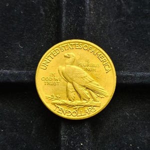 May include: A gold coin featuring an eagle design, with the inscriptions "United States of America", "In God We Trust", and "Ten Dollars". The coin is circular and shows signs of wear.