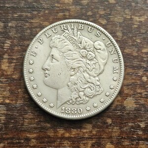 Puede incluir: Una moneda de plata con el perfil de una mujer, con las palabras "E PLURIBUS UNUM" y "LIBERTY" visibles. El año "1880" también se muestra. La moneda está sobre una superficie de madera.