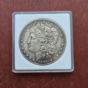 Puede incluir: Una moneda de plata de 1895, con el perfil de una mujer con cabello ondeado y las inscripciones "E PLURIBUS UNUM" y "LIBERTY". La moneda está dentro de un soporte de plástico transparente.