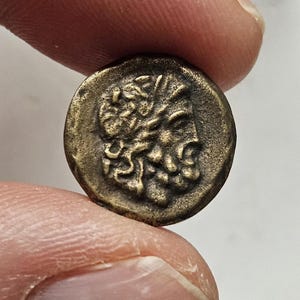 Peut inclure: Une petite pièce de monnaie ronde de couleur bronze, représentant un profil détaillé d'un homme barbu aux cheveux bouclés. La pièce est tenue entre deux doigts, mettant en valeur son design complexe et son aspect vieilli.