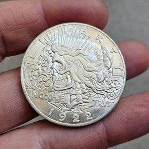 Puede incluir: Una moneda de plata con un diseño de calavera, con el texto "IN GOD WE TRUST" y el año "1922". La moneda tiene un relieve detallado con una calavera y cabello suelto, y se sostiene en una mano.