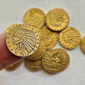 Puede incluir: Una colección de monedas de oro de cabeza de indio, con el perfil de un nativo americano con tocado. Una moneda se sostiene con un dedo, mostrando la fecha "1915" y la palabra "LIBERTY" inscrita en la moneda.