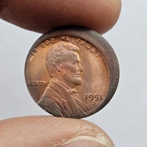 Puede incluir: Una moneda de Lincoln de 1951, de color cobre. El anverso presenta un perfil de Abraham Lincoln, con las inscripciones "IN GOD WE TRUST" y "LIBERTY". También es visible el año "1951".