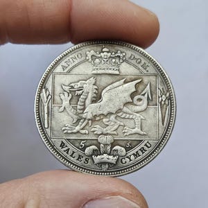 Puede incluir: Una moneda de plata con un diseño detallado de un dragón, con las palabras "WALES CYMRU" y "ANNO DOM". La moneda se sostiene entre los dedos, mostrando sus intrincados detalles.