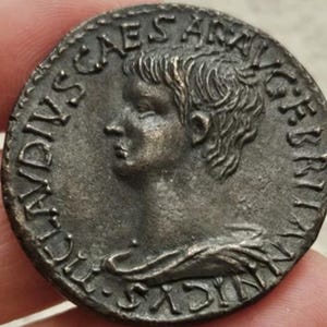Könnte beinhalten: Eine bronzene römische Münze mit einem Profil einer Person mit detaillierten Haaren und Gesichtszügen. Der Rand der Münze zeigt Inschriften, wobei die Wörter "CAESAR" und "BRIT" sichtbar sind. Die Münze hat ein verwittertes Aussehen.