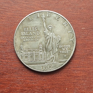 Puede incluir: Una moneda de plata con la Estatua de la Libertad sosteniendo una antorcha, con la palabra "LIBERTY" en la parte superior. La moneda también incluye el texto "ELLIS ISLAND GATEWAY TO AMERICA" y el año "1906".