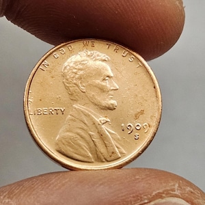 以下が含まれることがあります： 銅製の1909 Sリンカーンセント硬貨。表面にはアブラハム・リンカーンの横顔、単語「LIBERTY」、日付「1909」が描かれています。硬貨には「IN GOD WE TRUST」の文字も刻まれています。