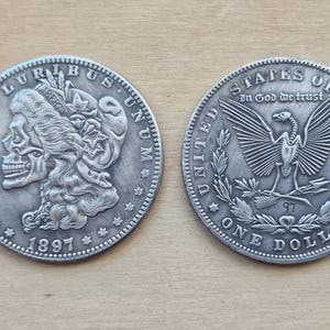 Puede incluir: Dos monedas de un dólar de plata, una con una calavera y la otra con un águila. La moneda con la calavera tiene la inscripción "E. Pluribus Unum" y "1897". La moneda con el águila tiene la inscripción "United States of America" y "In God We Trust".