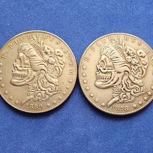 Puede incluir: Dos antiguas monedas de plata Morgan, cada una con un perfil detallado del rostro de una mujer, el año de acuñación es visible. Las monedas están sobre un fondo azul.