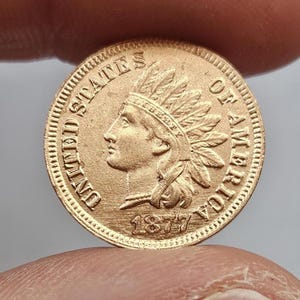 Puede incluir: Una moneda de oro de 1877, Indian Head. La moneda presenta un perfil de un nativo americano con un tocado de plumas y las palabras "UNITED STATES OF AMERICA". El año 1877 es visible debajo.