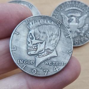 Puede incluir: Una moneda de un dólar de plata con un diseño de calavera y huesos cruzados. La moneda tiene las palabras "Liberty", "In God We Trust" y "1974".
