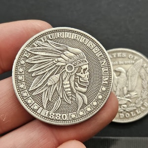 Puede incluir: Una moneda plateada con un grabado detallado de un nativo americano con un tocado y una calavera. La moneda tiene las palabras "PLURIBUS UNUM" y "1880" inscritas en ella.