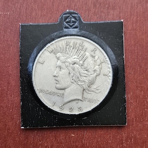 Puede incluir: Una moneda de plata de 1923 Peace Dollar, con un perfil de Lady Liberty, está encerrada en un soporte protector negro. Los detalles de la moneda, incluida la inscripción "IN GOD WE TRUST", son visibles. El soporte tiene una pequeña parte superior decorativa.