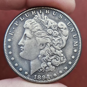 Puede incluir: Una moneda de plata con un perfil detallado de una mujer, la palabra "LIBERTY" sobre su cabeza y el año "1894" en la parte inferior. El borde de la moneda presenta pequeñas estrellas. Las palabras "E PLURIBUS UNUM" están inscritas alrededor del borde.