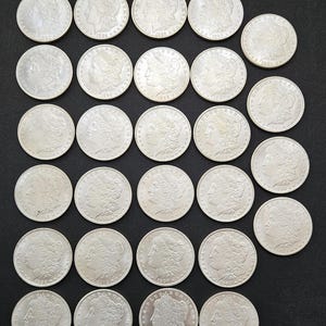 Puede incluir: Una colección de 30 monedas de plata Morgan dollar, cada una con un perfil de Lady Liberty en un lado y un águila en el otro. Las monedas están dispuestas en filas sobre una superficie negra.