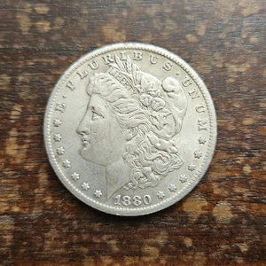 Puede incluir: Una moneda de plata de 1880, con el perfil de una mujer con el pelo suelto y la inscripción "LIBERTY". La moneda está rodeada de estrellas y las palabras "E PLURIBUS UNUM".