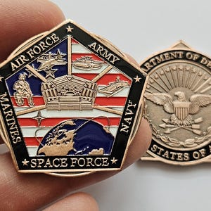 May include: A gold-colored pentagon-shaped challenge coin with the seals of the United States Armed Forces. The front features the seals of the Air Force, Marines, Army, Navy, and Space Force, with a globe in the center. The back features the Department of Defense seal with the words "Department of Defense" and "United States of America".