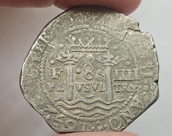 May include: A silver coin with an irregular shape, held between fingers. The coin features intricate engravings, including a crown, pillars, and the number 8. The coin's surface shows signs of wear and age, with visible text and symbols.