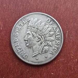 Puede incluir: Una moneda de plata que representa el perfil de un nativo americano con un tocado de plumas. La moneda está inscrita con "UNITED STATES OF AMERICA" y "LIBERTY". La moneda tiene un aspecto desgastado y antiguo.