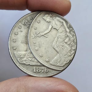 Könnte beinhalten: Zwei Silbermünzen, von denen sich eine teilweise überlappt. Die Münzen zeigen ein detailliertes Relief einer Frau, die einen Zweig hält, umgeben von Sternen. Das Jahr 1876 ist auf einer Münze zu sehen. Das Wort "LIBERTY" ist ebenfalls sichtbar.