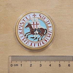May include: A circular emblem with a gold border, featuring a light blue and white design. It depicts two figures on a brown horse, with red crosses on their shields. The text "SIGILLVM MILITVM XPISTI" is around the edge.
