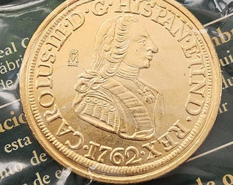 Könnte beinhalten: Eine goldfarbene Münze mit dem Profil eines Mannes mit gepuderter Perücke und der Inschrift "CAROLUS III D. G. HISPAN. ET IND. REX. 1762 XF". Die Münze befindet sich in einer Schutzhülle aus Kunststoff, mit Text von "EL PAIS".