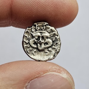 Può includere: Una piccola moneta color argento con un design in rilievo dettagliato. La superficie della moneta ha un aspetto strutturato e invecchiato, e il disegno centrale sembra raffigurare un volto o una figura stilizzata. La moneta è tenuta tra due dita.