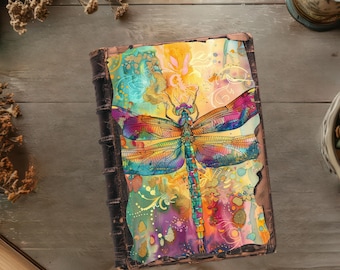May include: A colorful dragonfly with intricate details is painted on the cover of a leather-bound journal. The background features a watercolor wash of yellow, orange, pink, and purple.