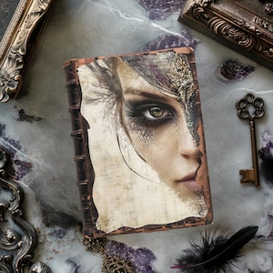 May include: A vintage-style leather-bound journal with a detailed illustration of a woman's face on the cover. The woman has a golden eye and a textured, metallic design on her face.